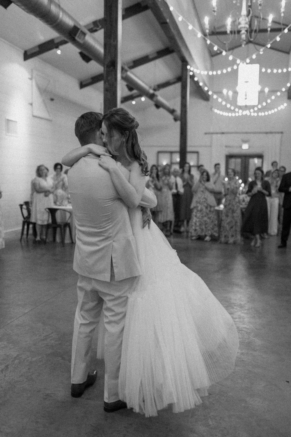 black and white image of a bride and groom dancing. he is lifting her up as she hugs him, smiling. loved ones watching