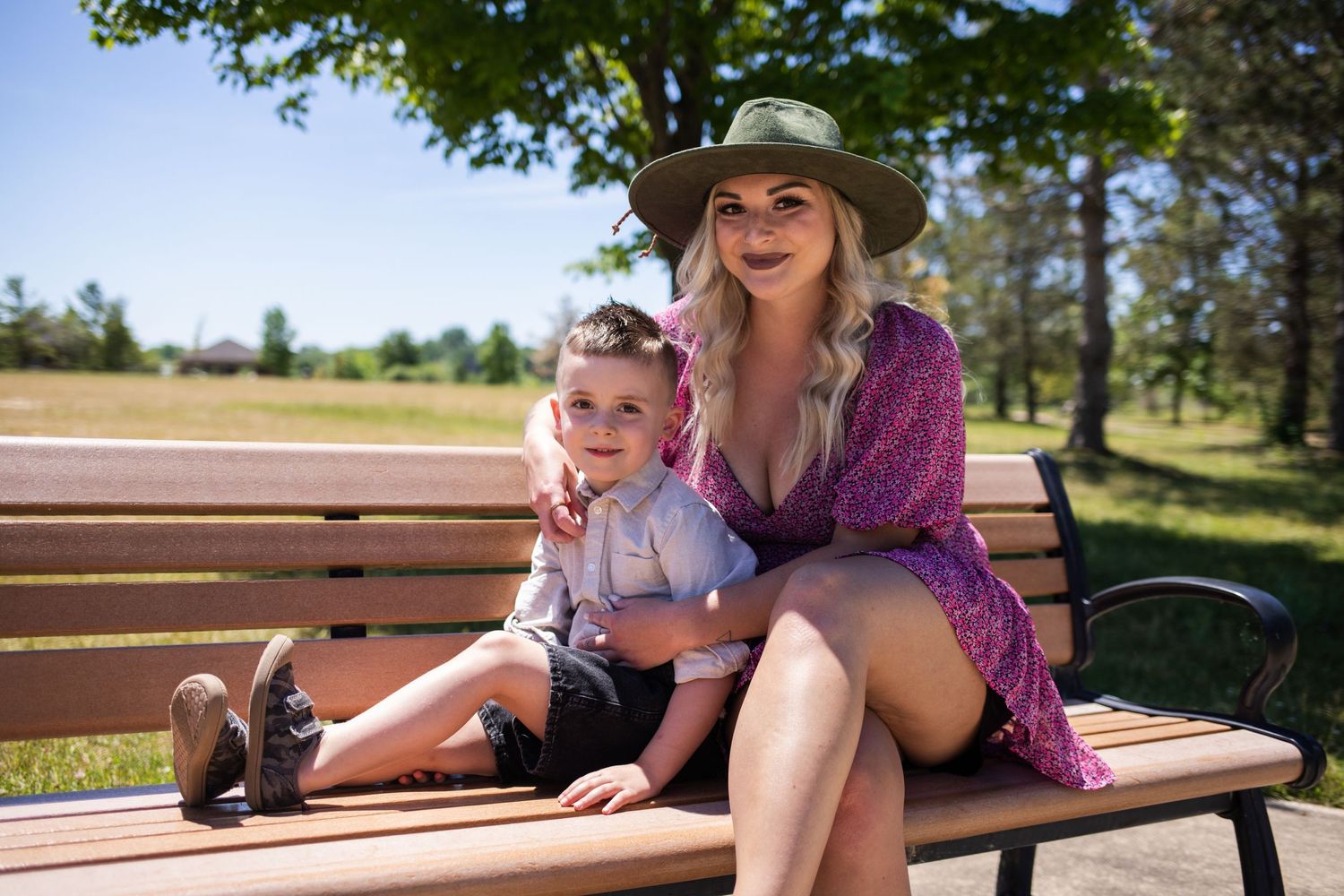 Blonde woman in floral dress and hat sitting on a park bench with a young toddler.