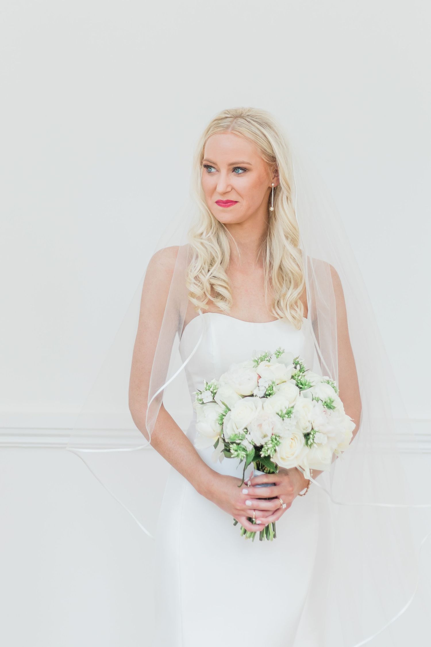 Minimalist bridal portrait against white wall featuring elegant bouquet and fitted dress.