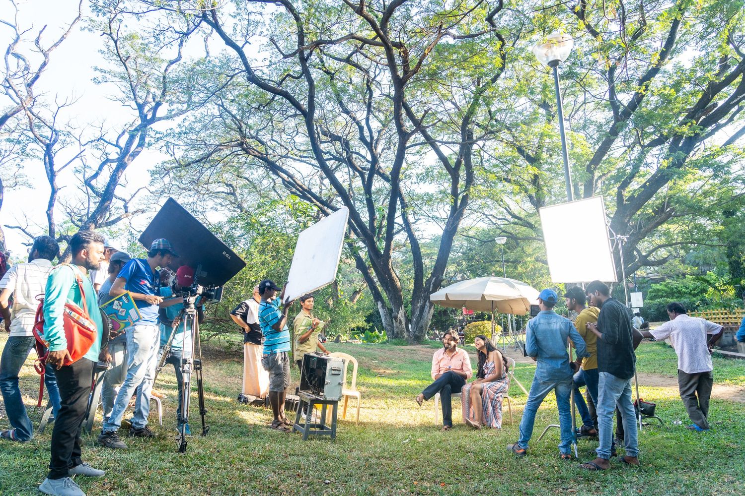 Film crew and production team setting up equipment under trees for an outdoor shoot.
