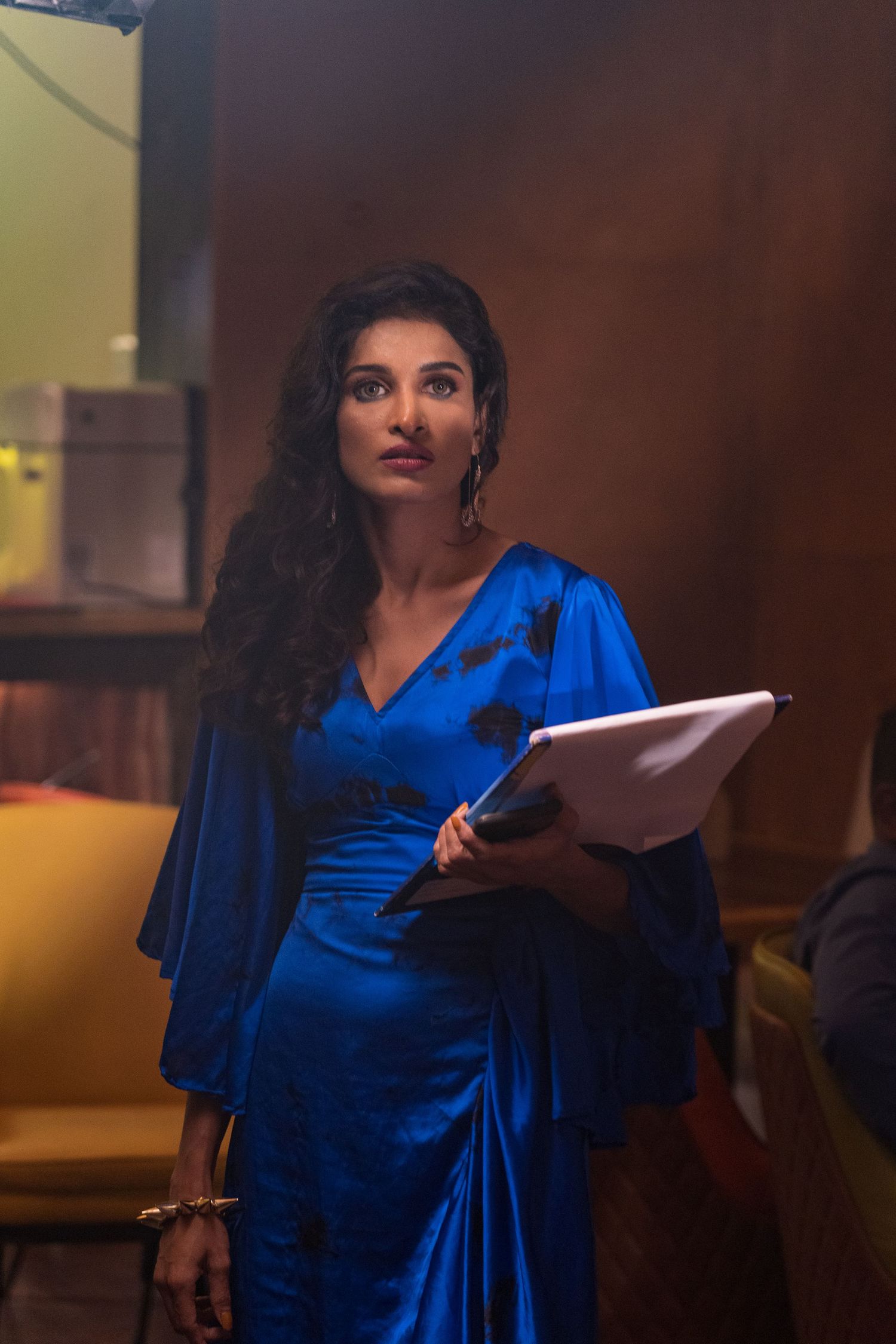 A woman in a brilliant blue dress stands thoughtfully while holding documents in a warmly lit setting.