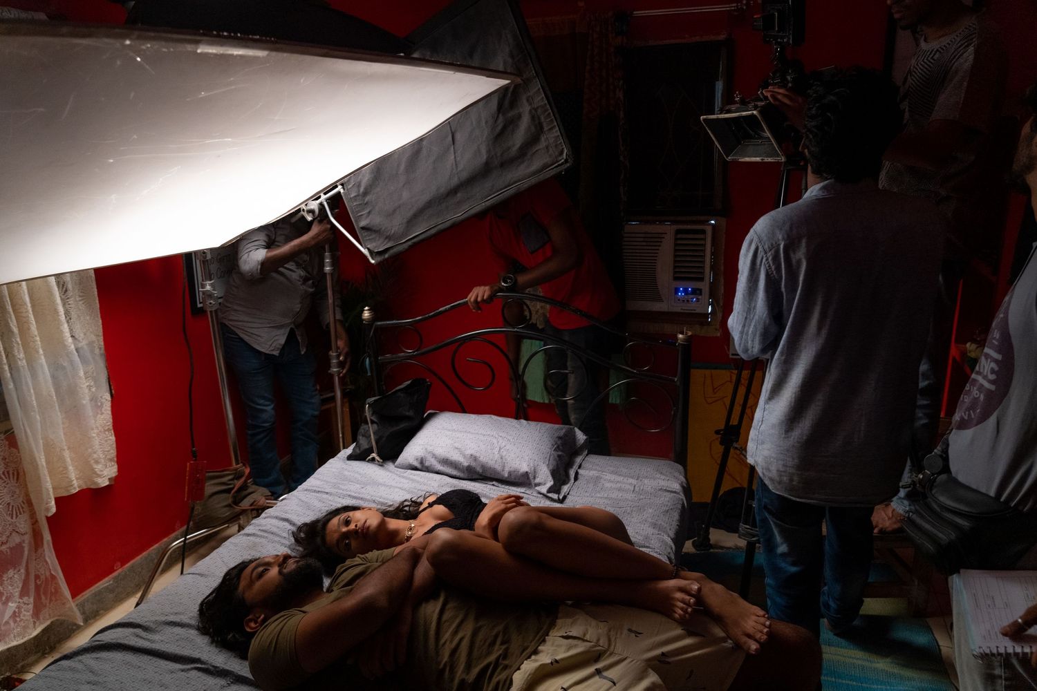 Film crew sets up lighting and camera equipment in a red-walled room for a bedroom scene.