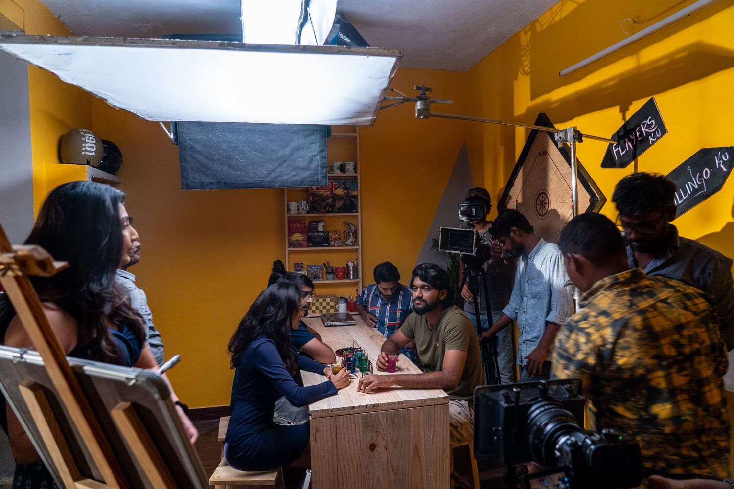 Production team films around a wooden table in a yellow-walled room with overhead lighting setup.