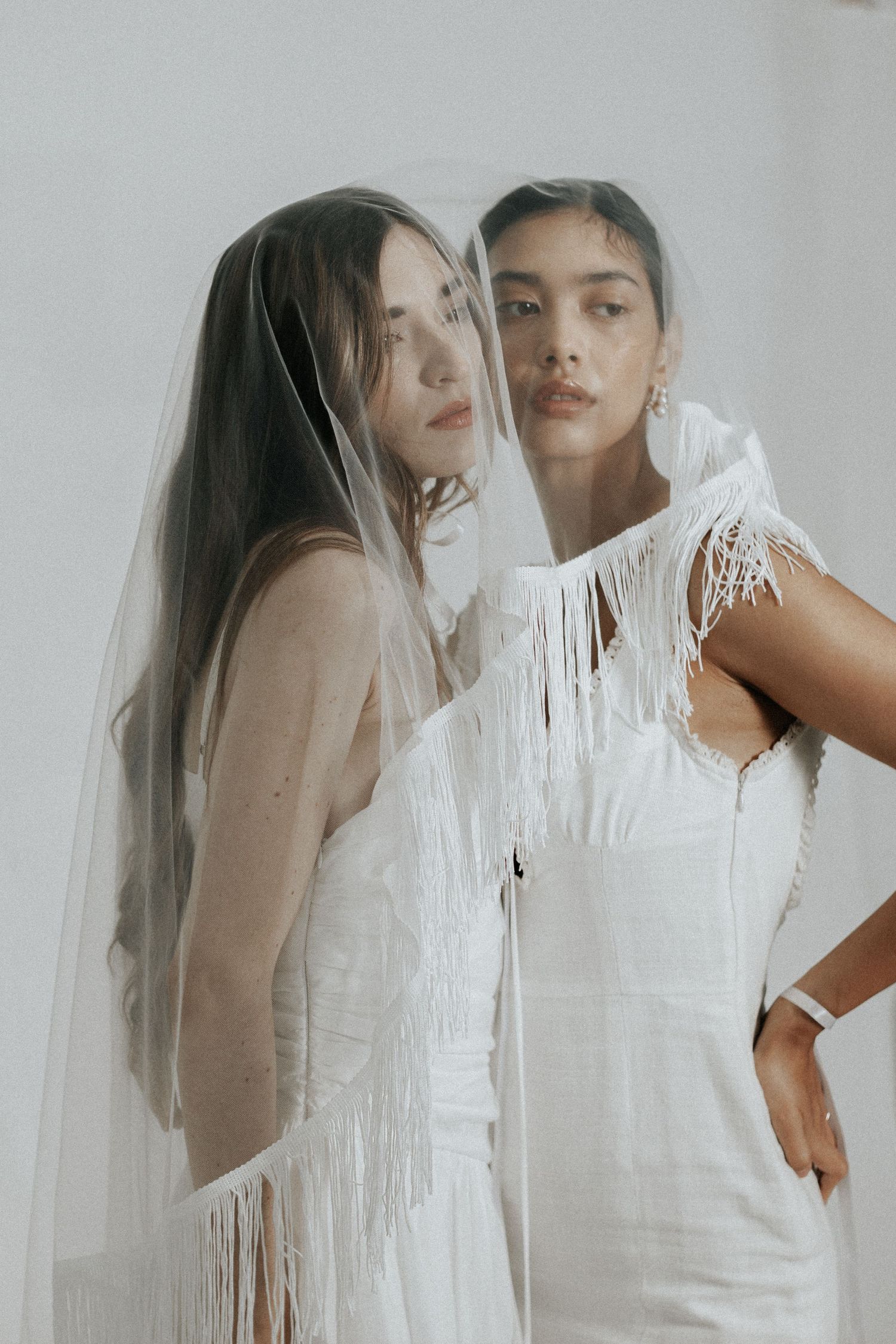 Two models in white dresses pose together with flowing veils in an ethereal fashion photo.