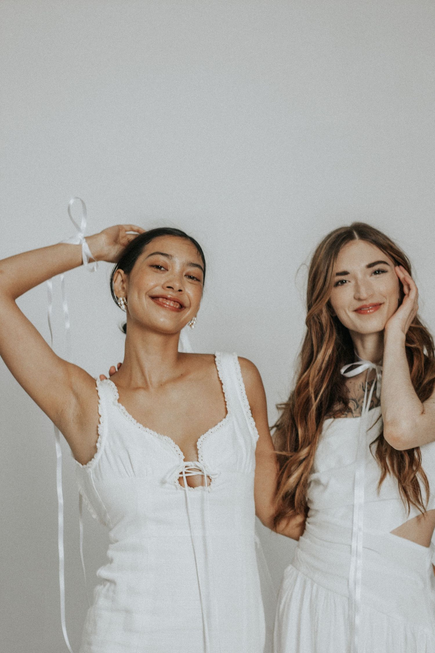 Two women in white dresses pose playfully against plain background.