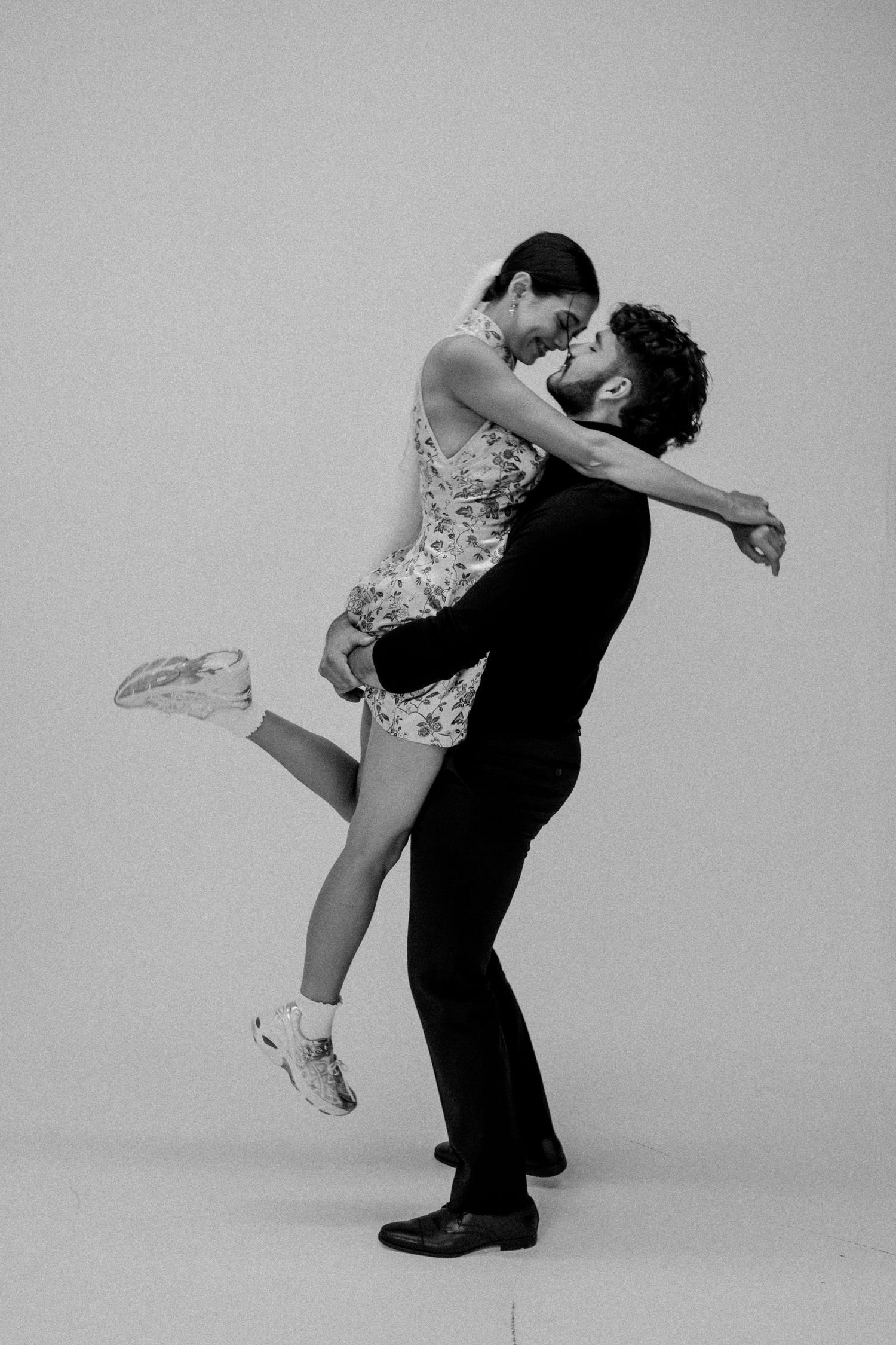 A couple in contrasting formal wear shares a playful dance pose against a light background.