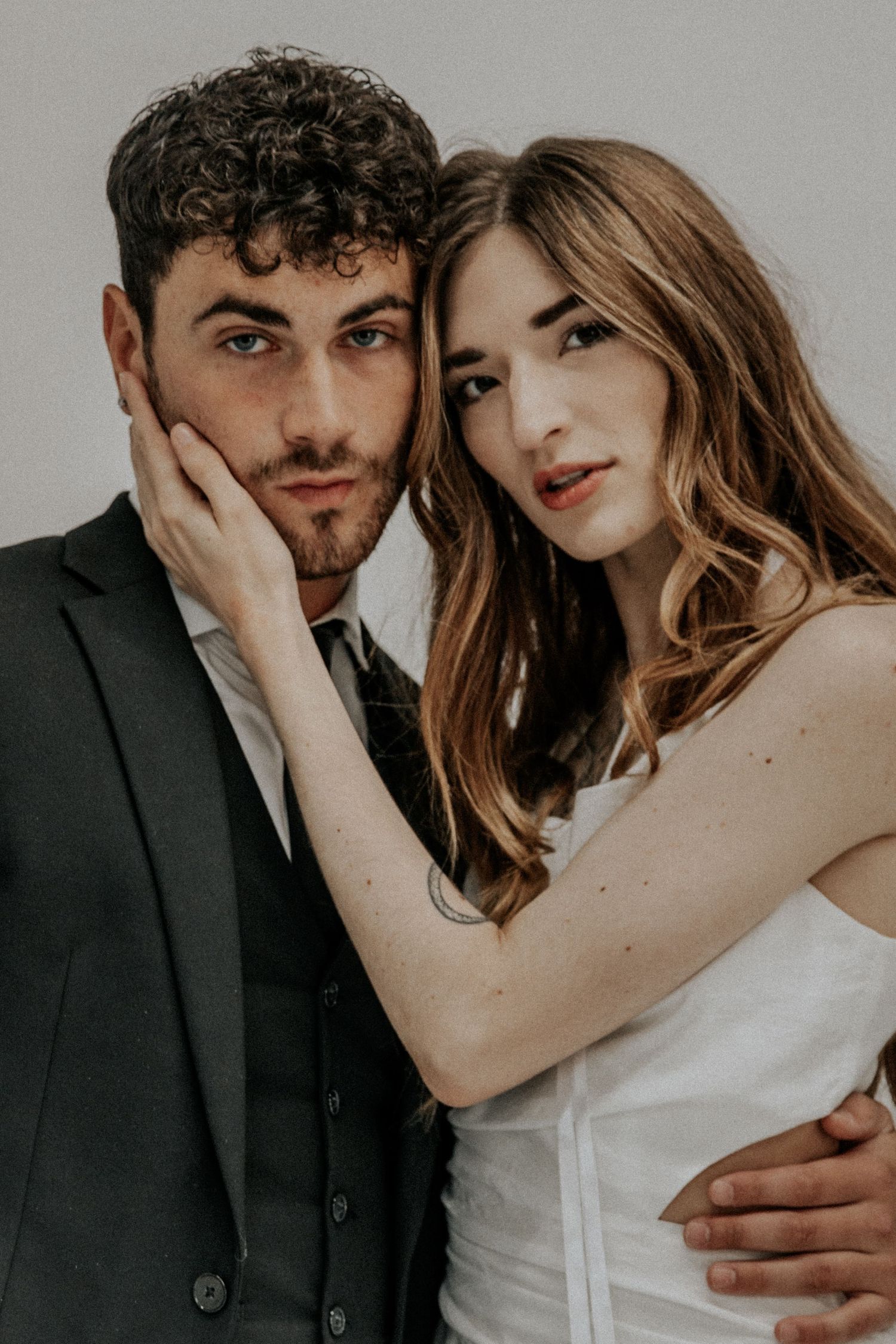 Stylish couple poses intimately against neutral background in wedding attire.