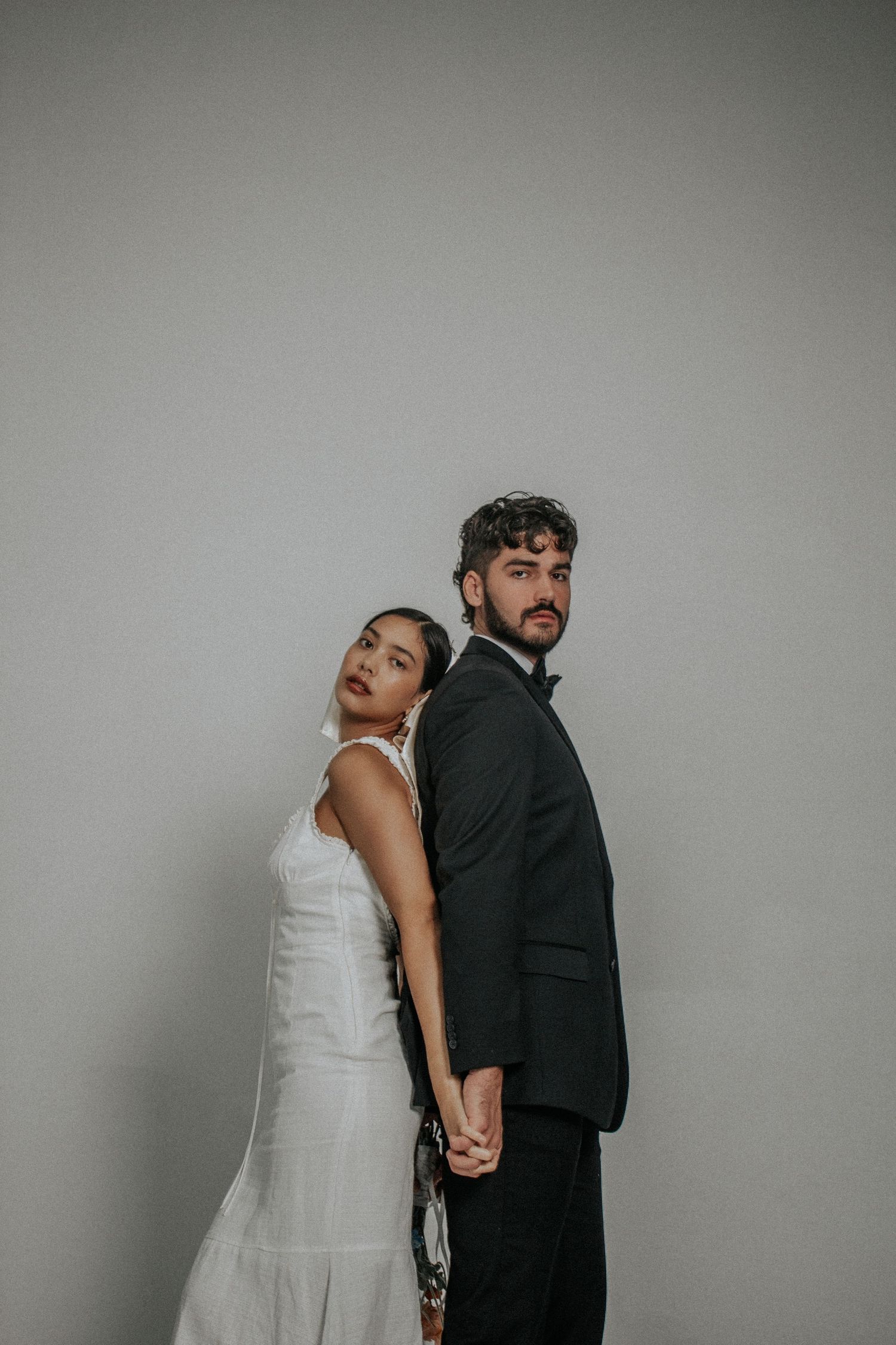A stylish couple in wedding attire poses back-to-back against a gray wall in a modern minimalist setting.