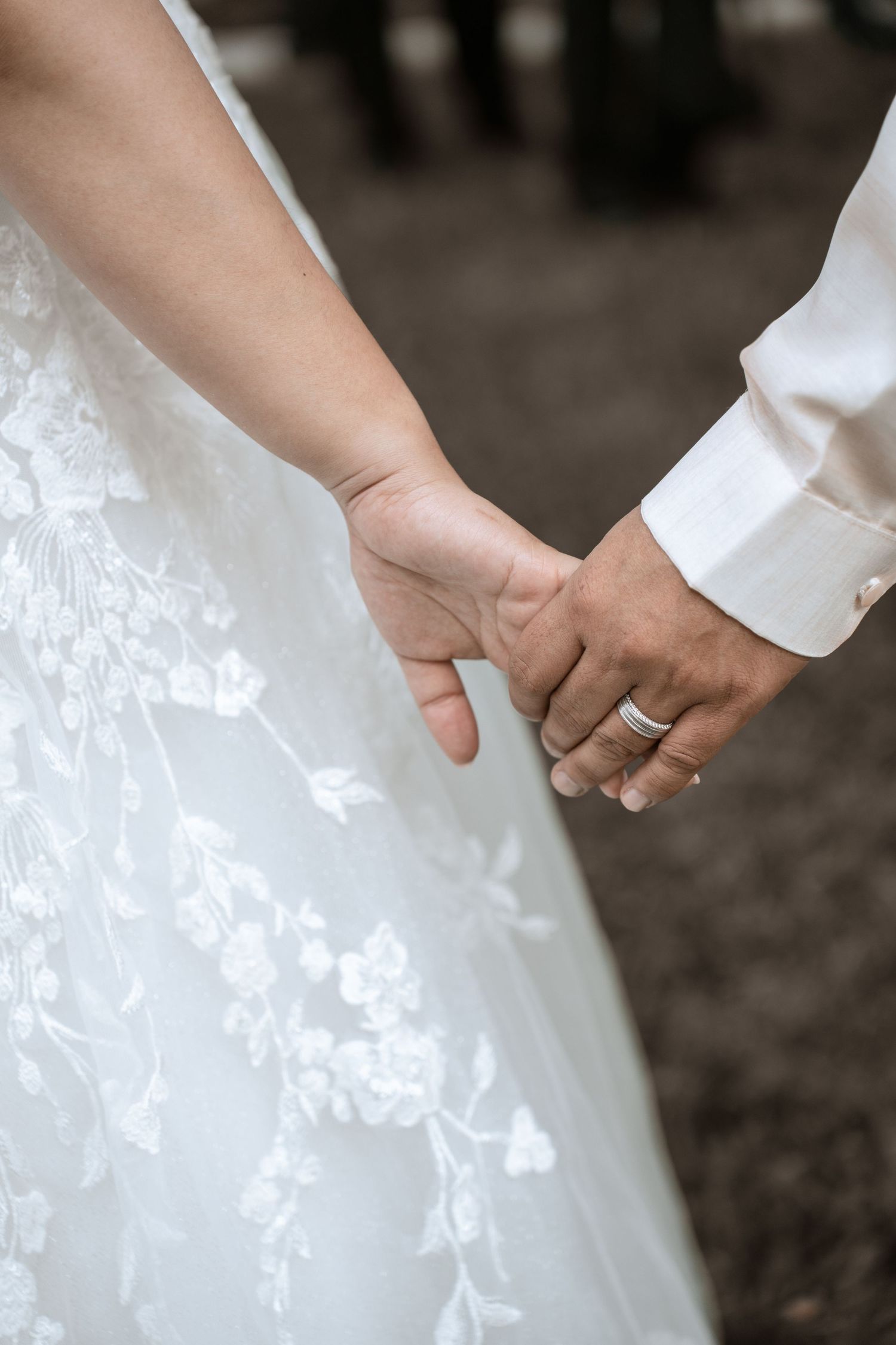 Close-up of newlyweds holding hands with wedding rings showing, bride wearing white lace dress.