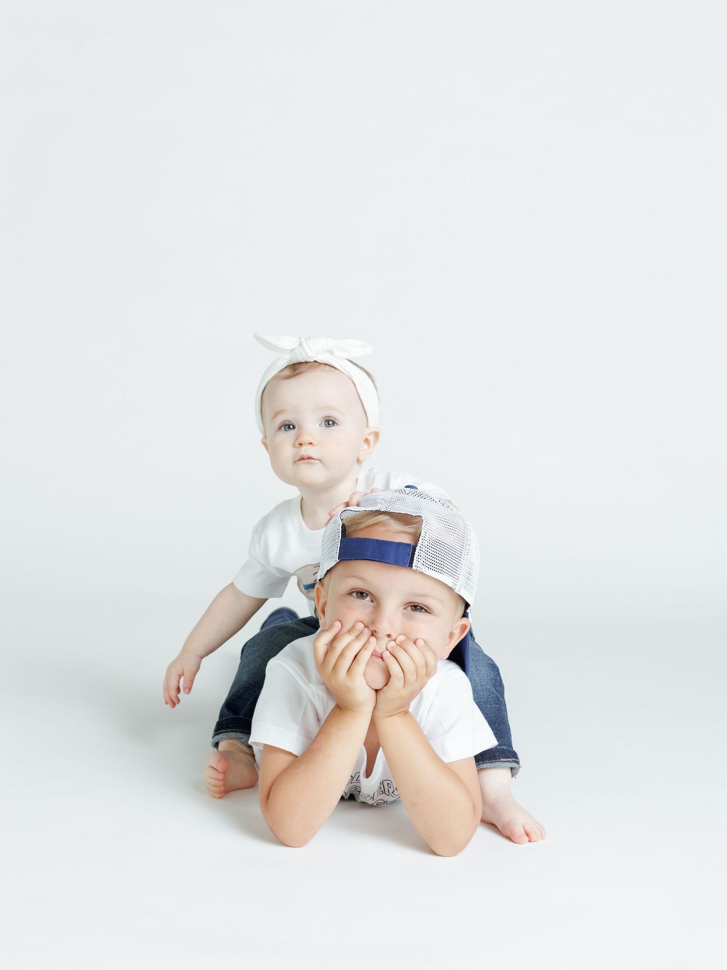 Two children in white clothing posing playfully against a minimal white background.