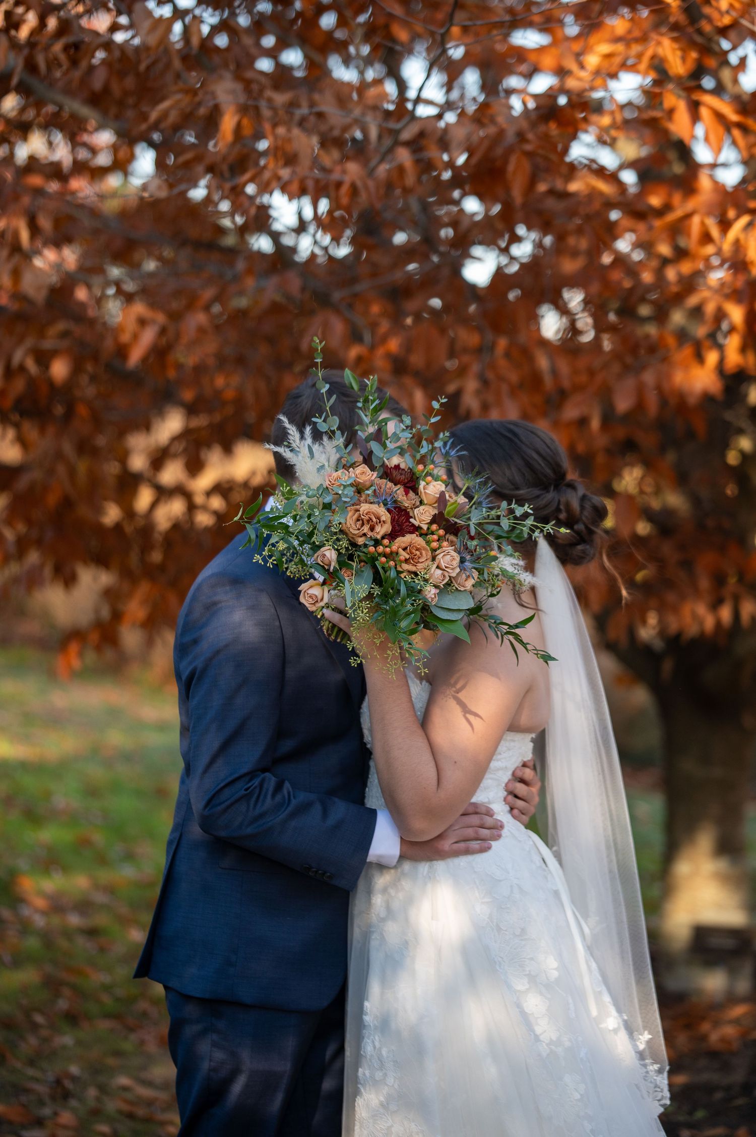 Romantic wedding kiss behind beautiful fall bouquet with orange autumn leaves in background.