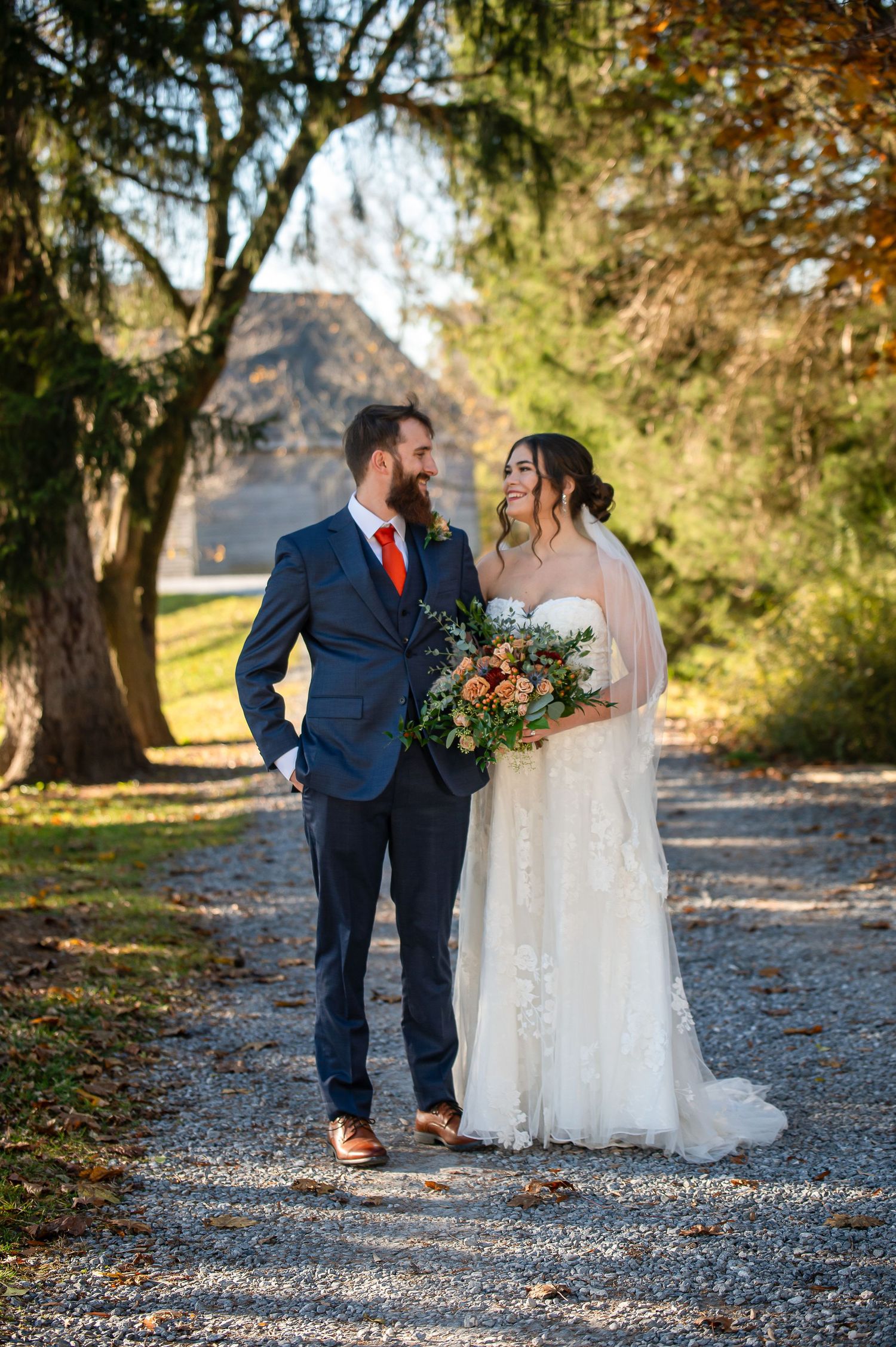 Wedding couple poses on gravel path with rustic barn and autumn foliage in background.