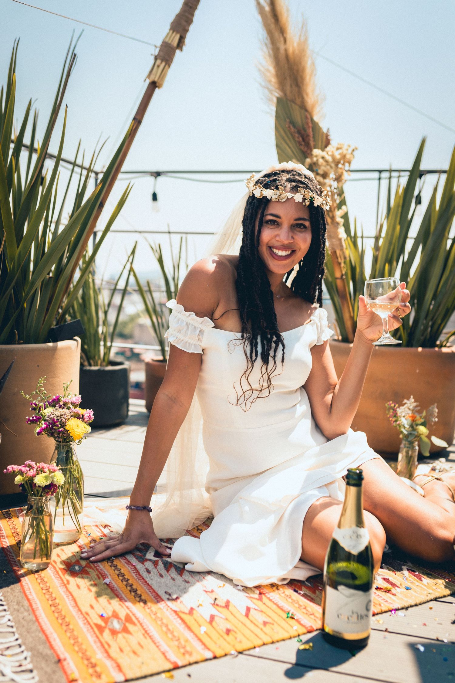 Boho-style picnic setup with white dress, floral crown, and champagne on patterned blanket.