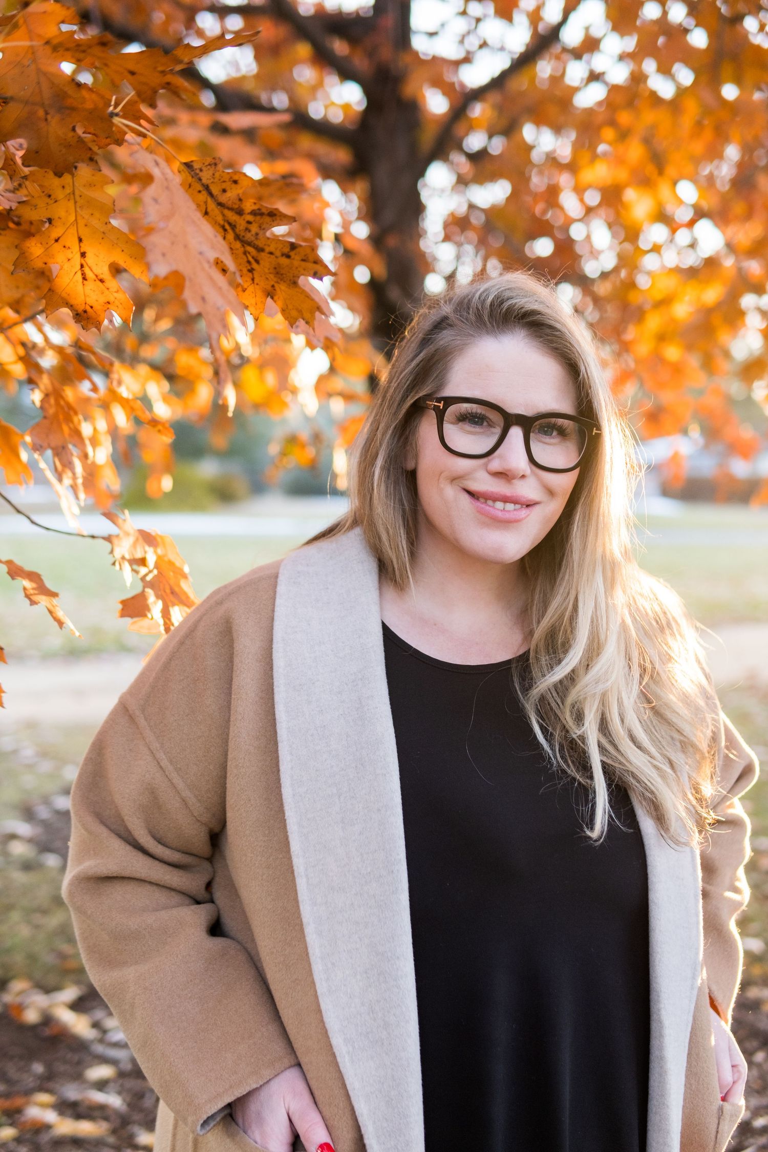 A woman wearing glasses and a brown coat smiles outdoors against colorful autumn foliage in golden light.