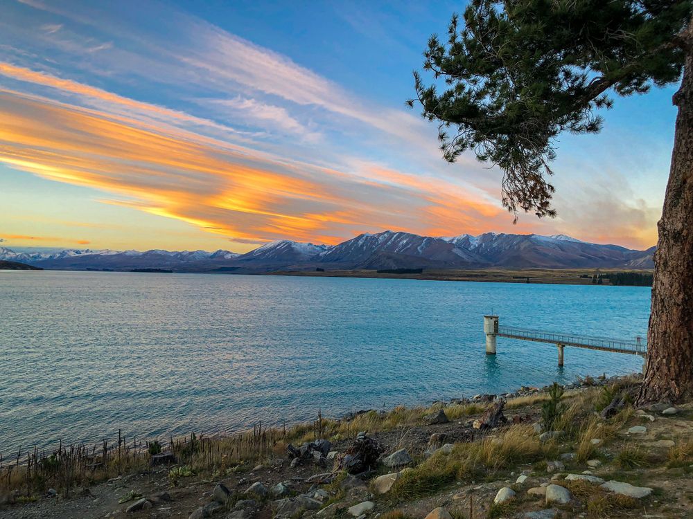 Lakeside sunset view with mountains and colorful sky reflected in calm waters.