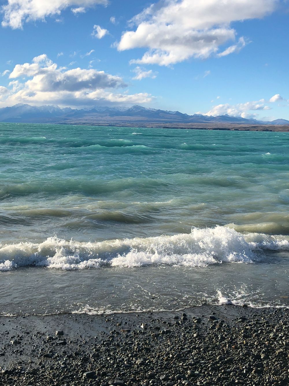 Rolling waves meeting the black pebble beach with mountains visible across the turquoise waters.