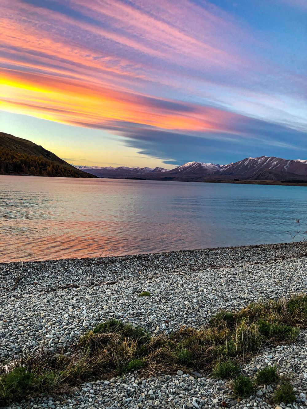 Rocky shoreline of Lake Tekapo at sunset with dramatic pink and orange clouds illuminating the mountain landscape.