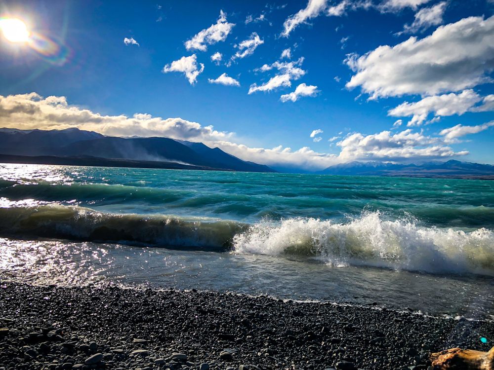 Turquoise waves crash against the rocky beach shore under bright blue skies with white puffy clouds.