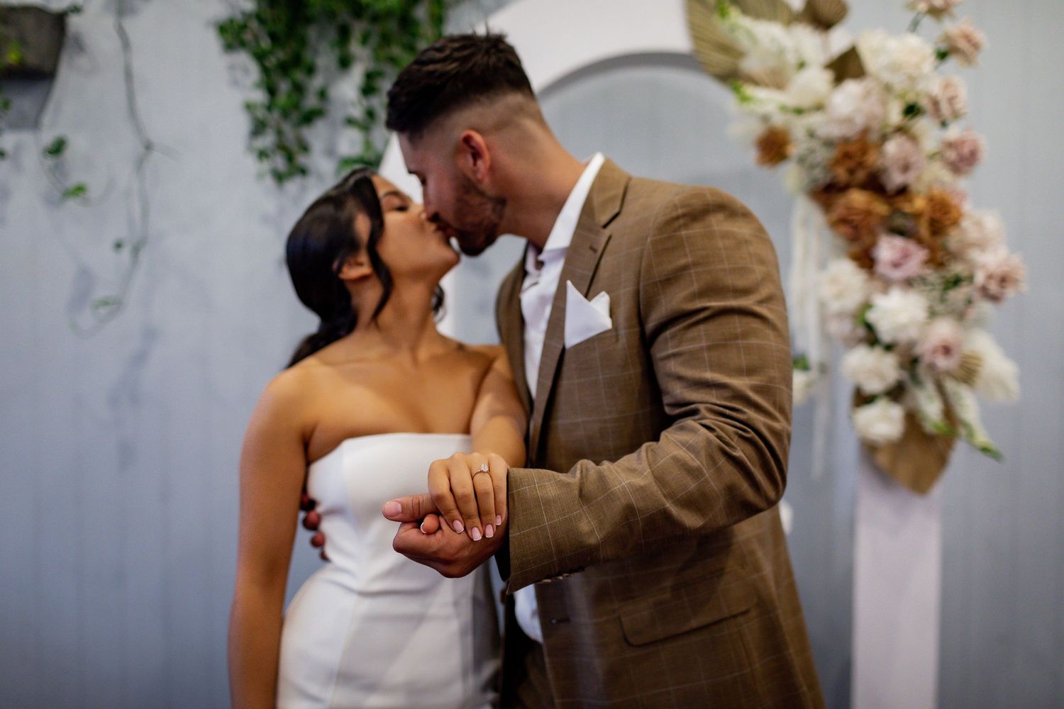 Romantic wedding couple sharing an intimate kiss against a white wall with floral decorations.