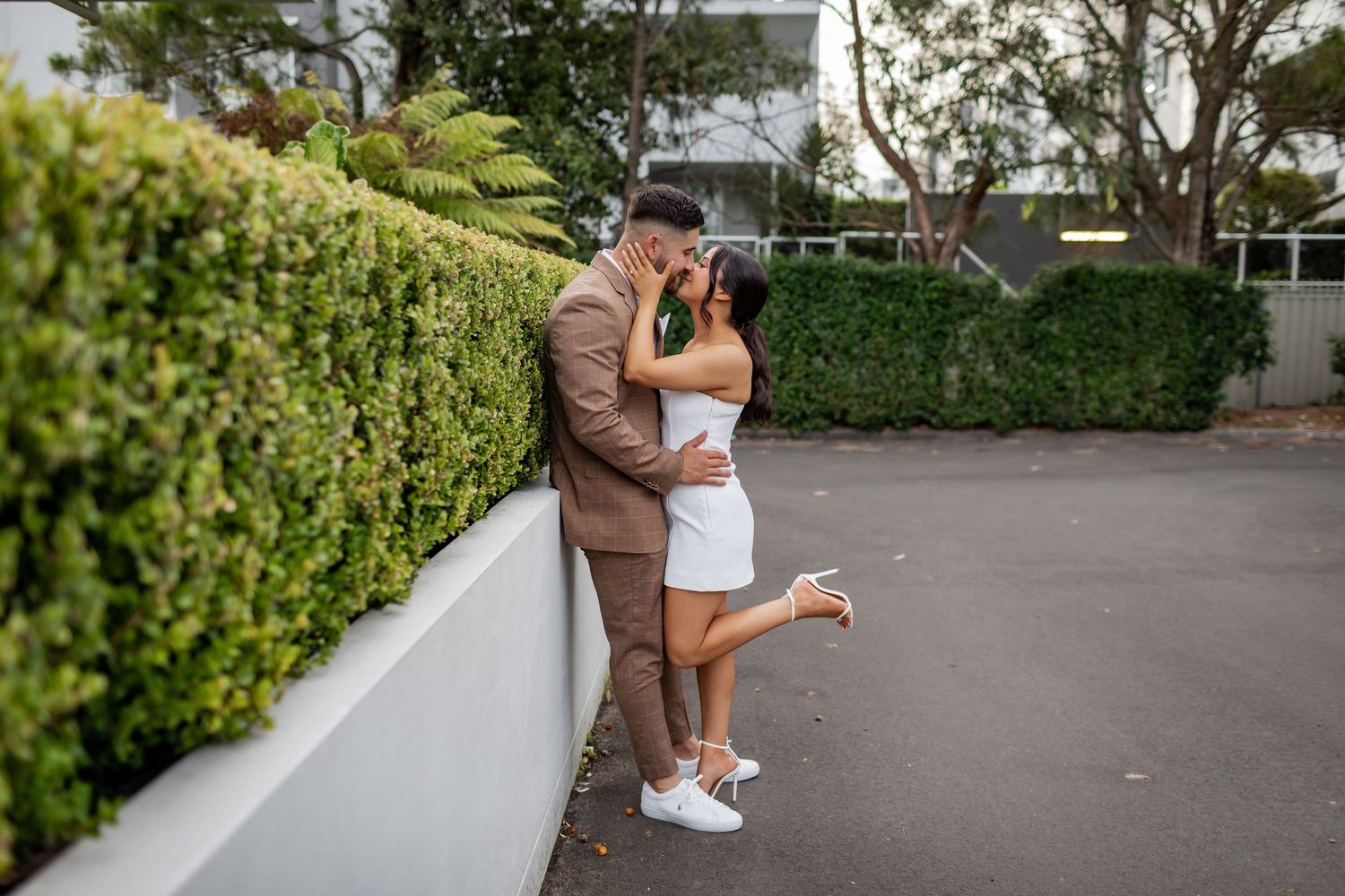 Stylish couple in formal attire sharing a tender moment against a garden wall.