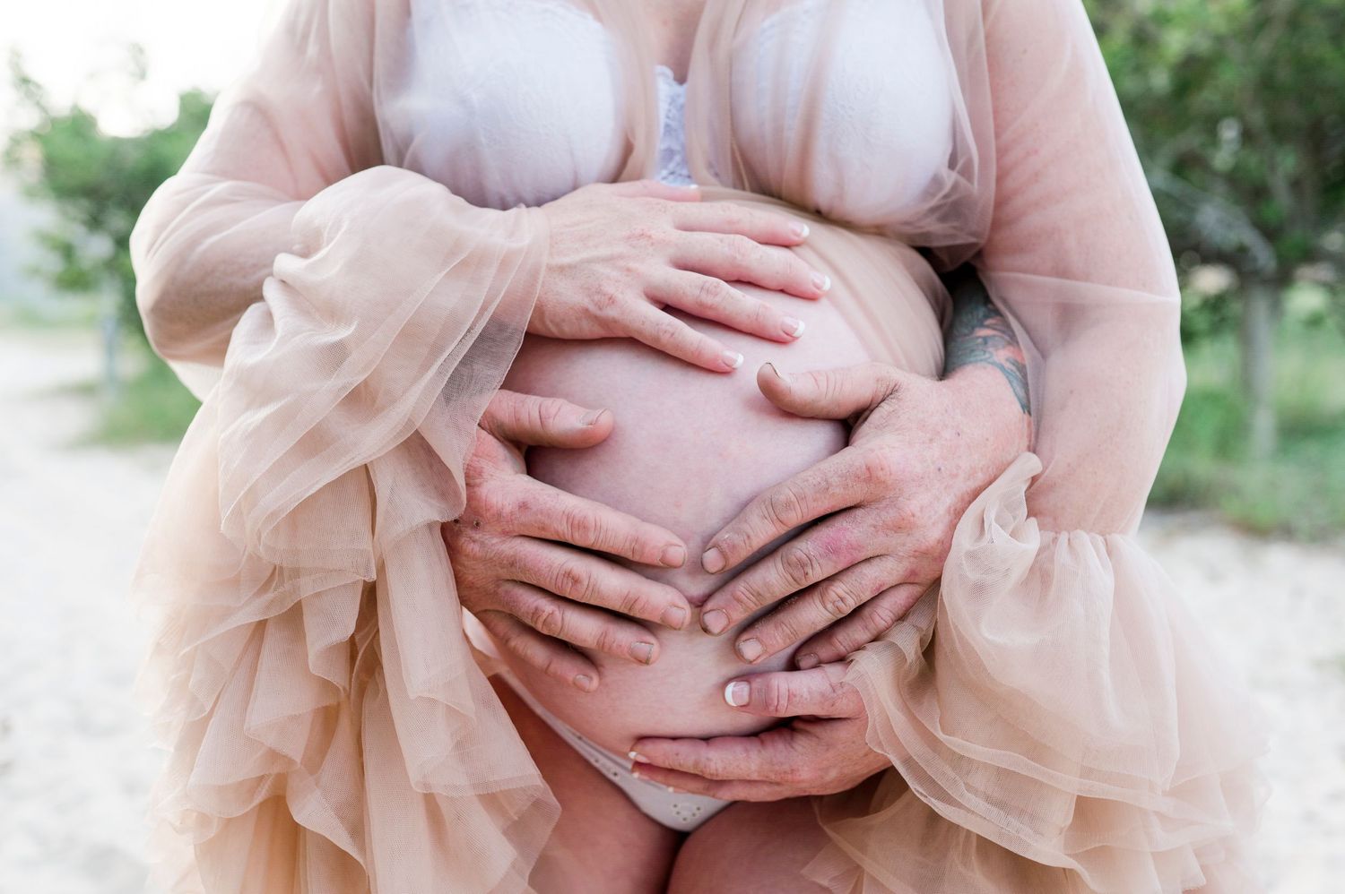 Hands cradle a pregnant belly in a flowing sheer pink dress against a natural outdoor backdrop.