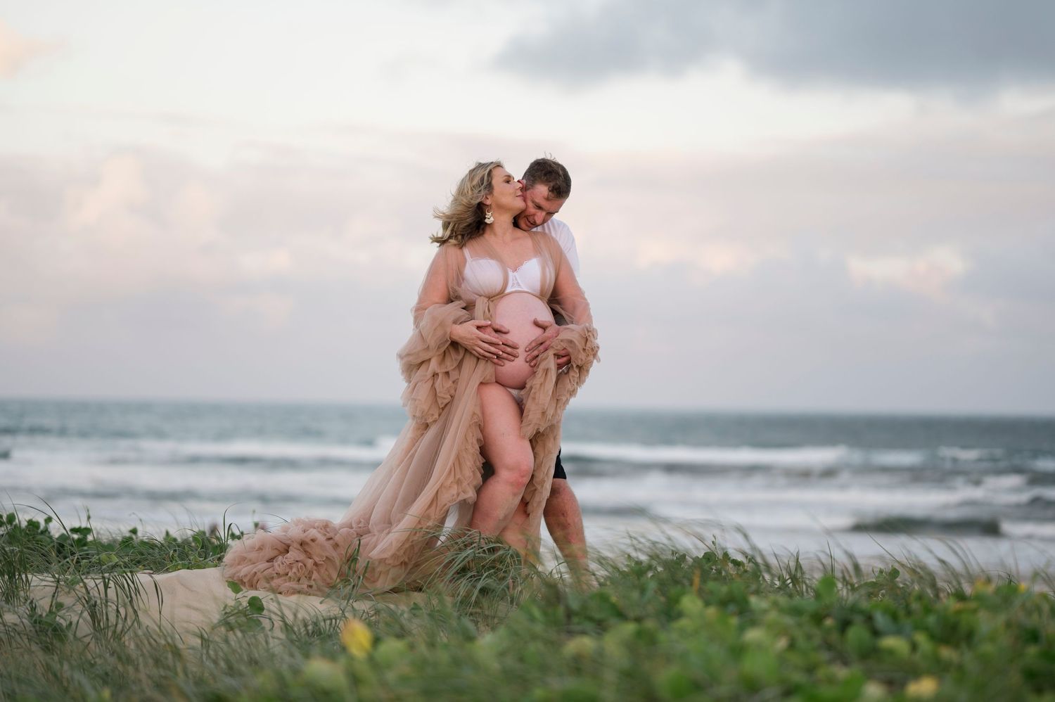 A romantic beach maternity photo with flowing blush dress against ocean backdrop.