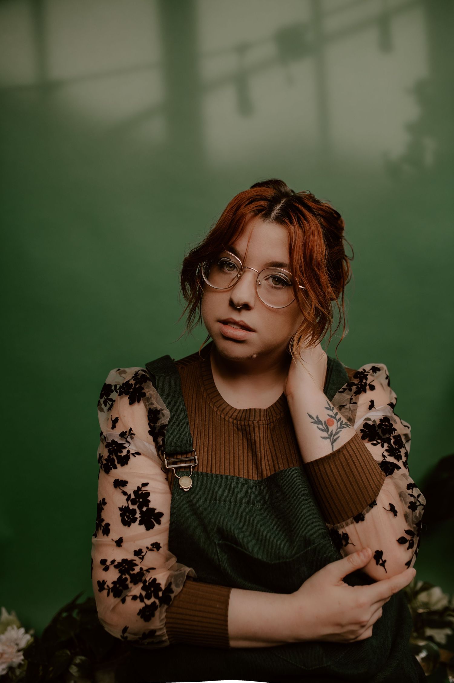 A portrait in front of a green background featuring sheer floral sleeves and a brown sweater.