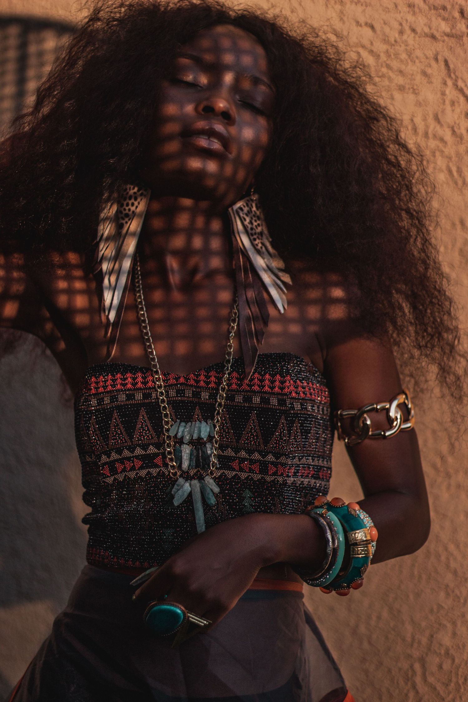 Artistic portrait with patterned light shadows and ethnic jewelry against terracotta wall.