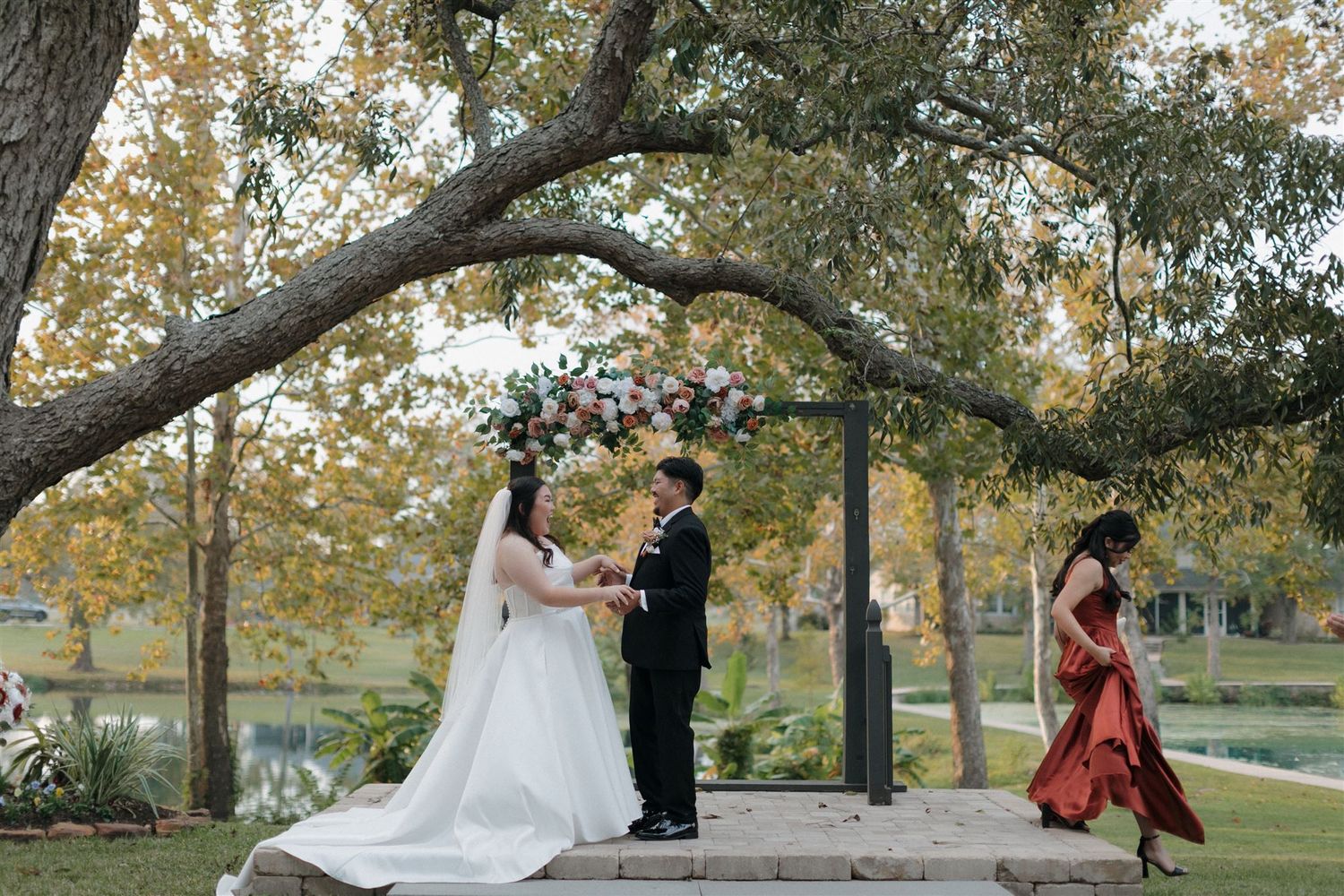 Wedding ceremony takes place on a wooden deck under large trees decorated with floral arrangements.