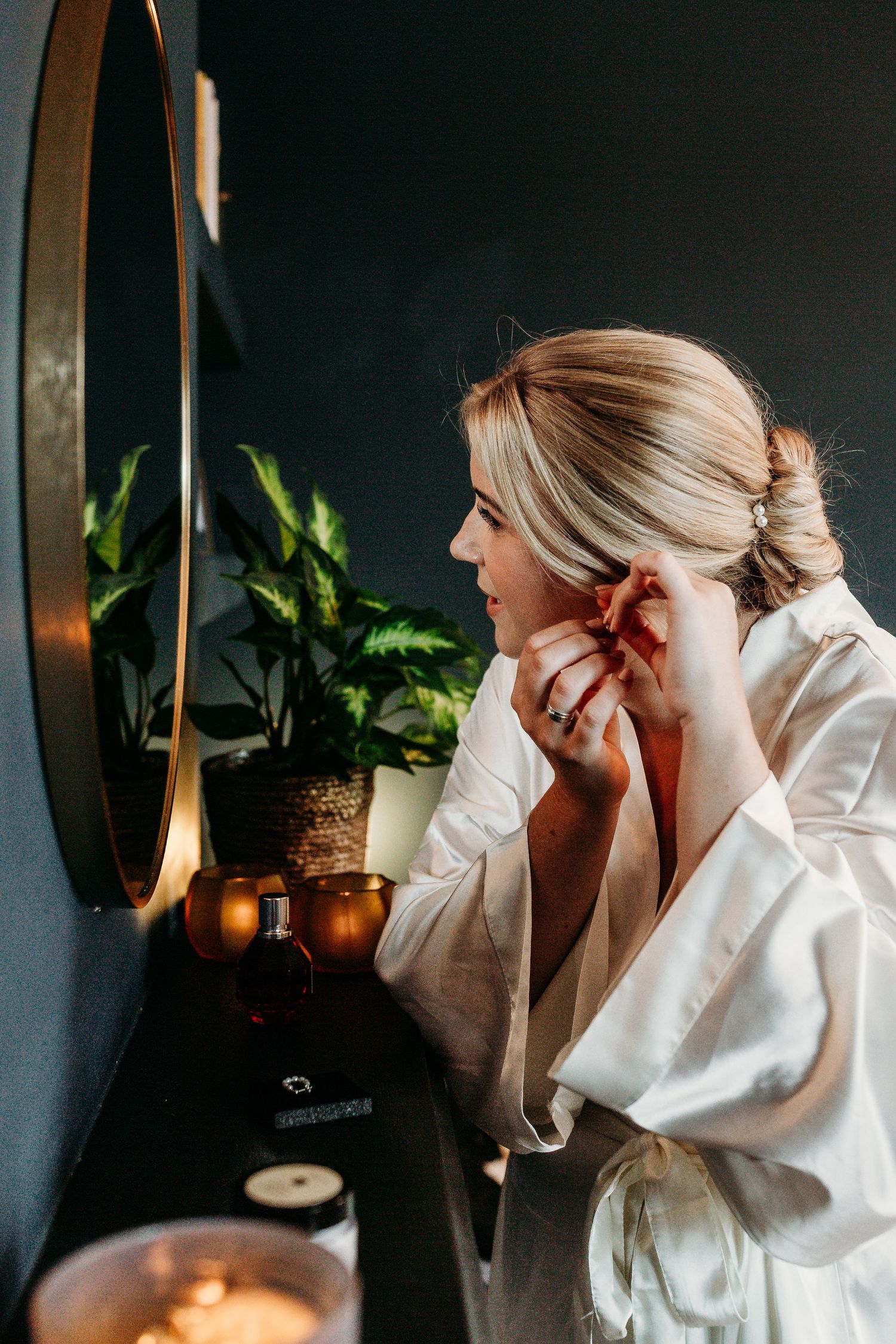 Woman in white robe putting on jewelry at a vanity table with lit candles and plants in the background.