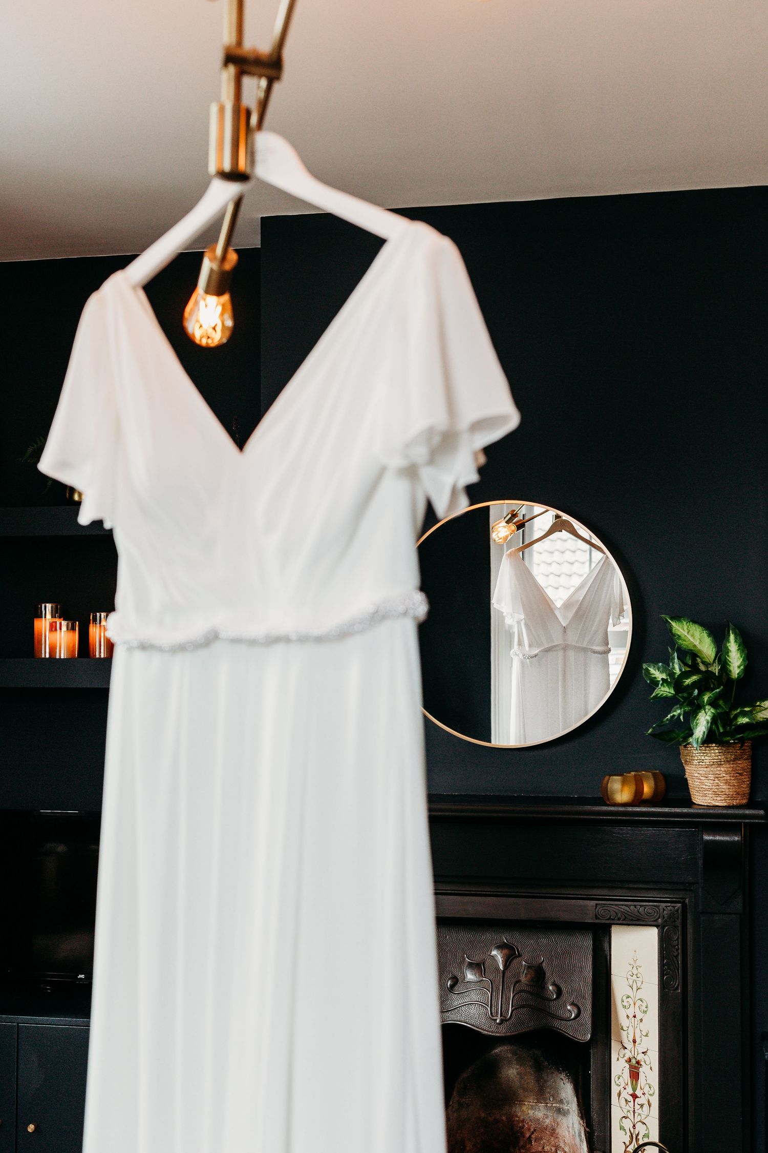 Elegant white wedding dress hanging against dark wall with round mirror and pendant light.