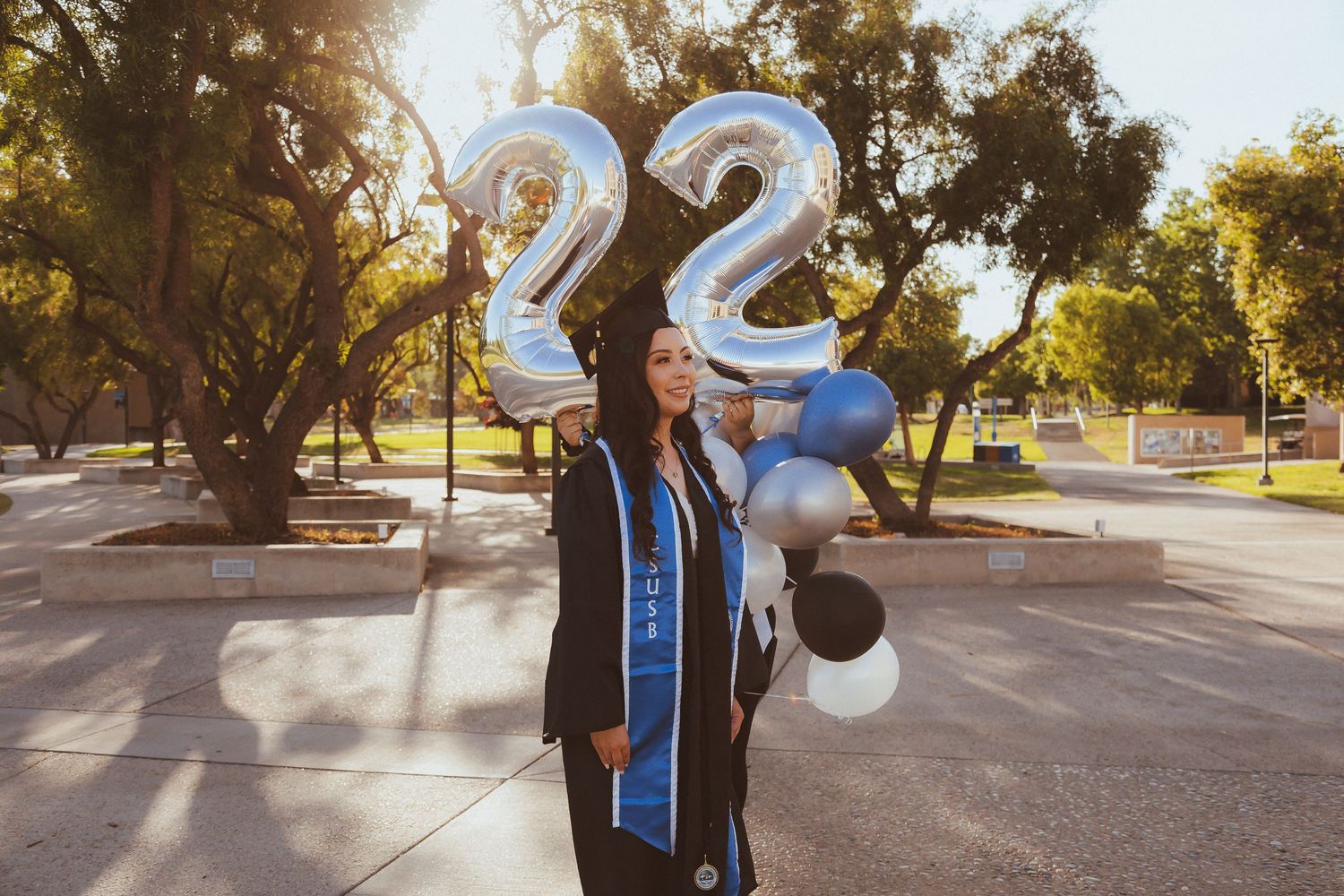Graduate celebrating with silver number 22 balloons and graduation sash in a sunny park setting.
