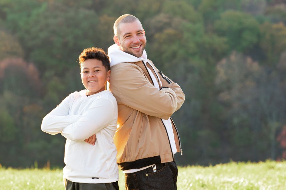 Father and son posing back to back in a sunny field with autumn trees in the background.