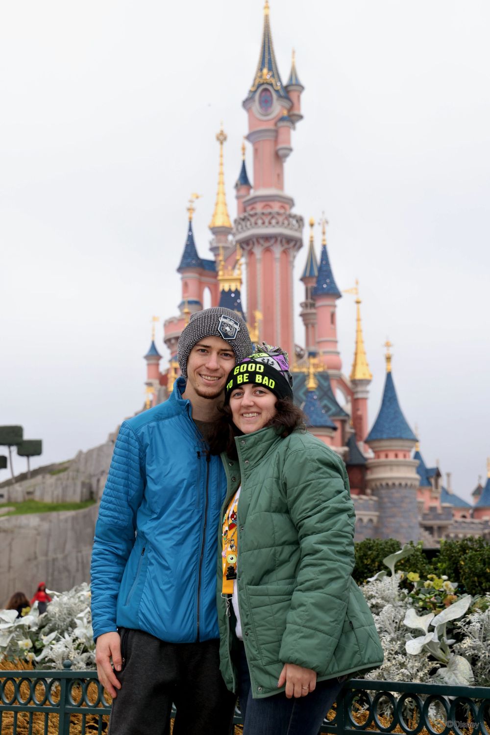 Couple posing together in front of Disneyland's iconic pink castle on a cloudy day.