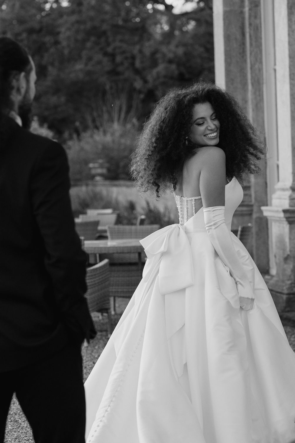 A bride in a stunning white ball gown with voluminous curls glances back playfully during a garden wedding photoshoot.