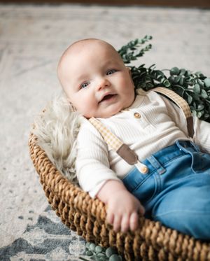Smiling baby wearing overalls lies in woven basket with eucalyptus sprigs.