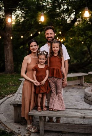 A family in coordinating rust-colored outfits poses on a wooden bench under string lights at dusk.