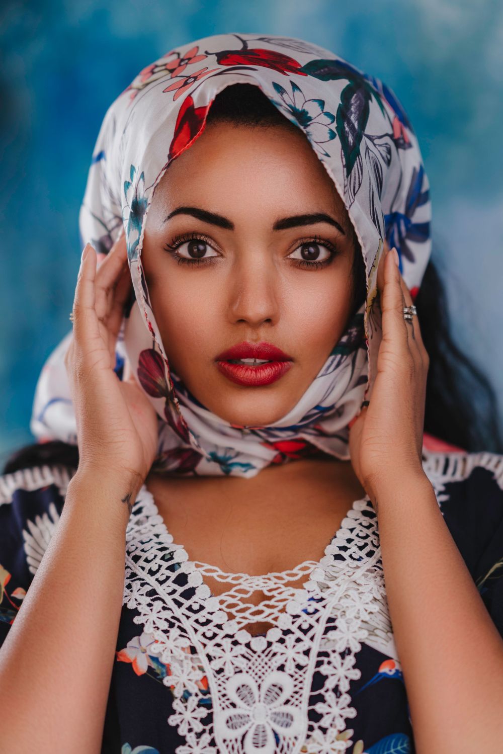 Portrait in floral headscarf and white lace top against blue background.