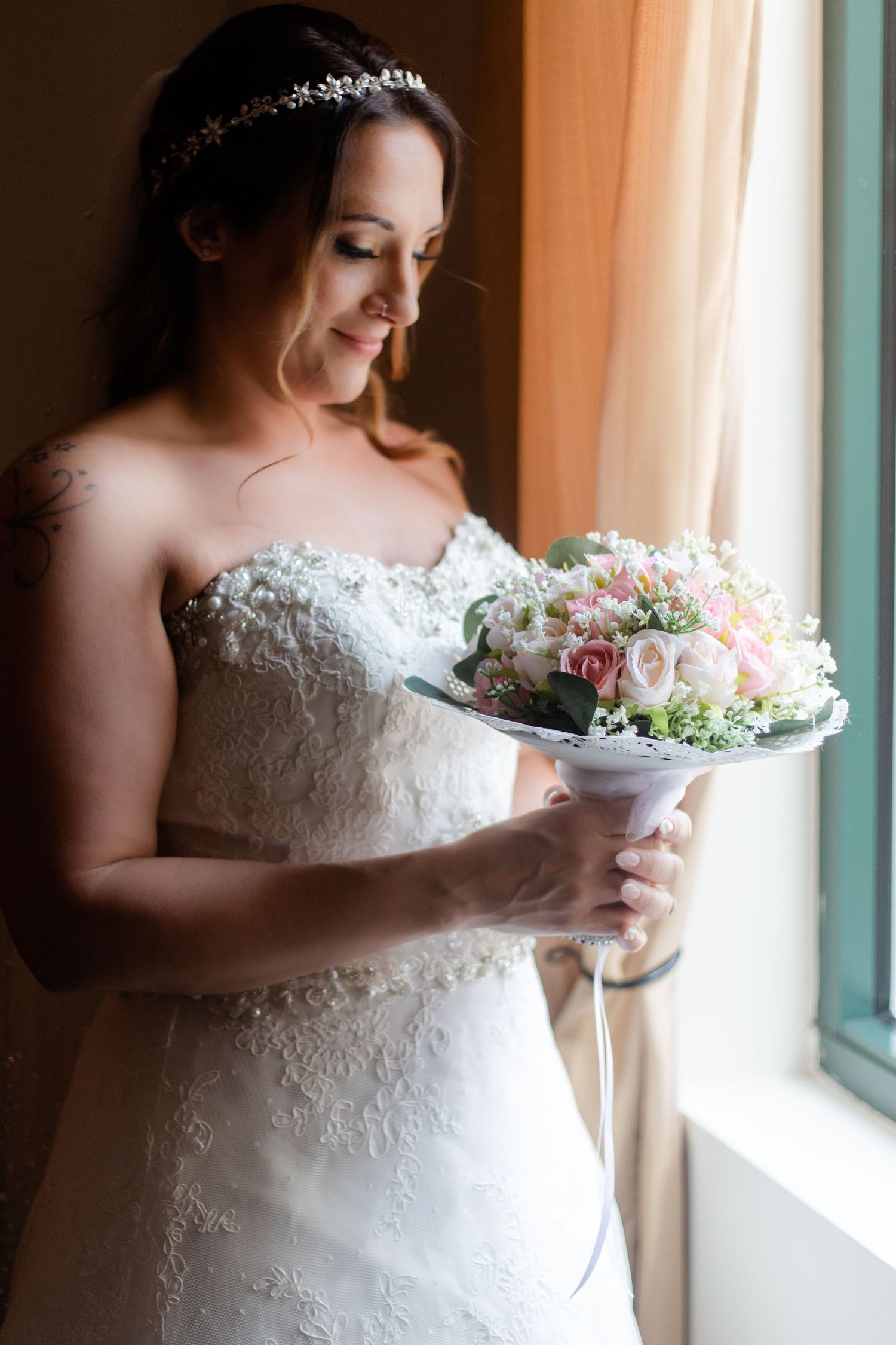 Bride in lace wedding dress holding a white floral bouquet near a window.