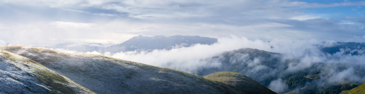 A panoramic mountain landscape with rolling peaks and valleys shrouded in dramatic clouds against a blue sky.