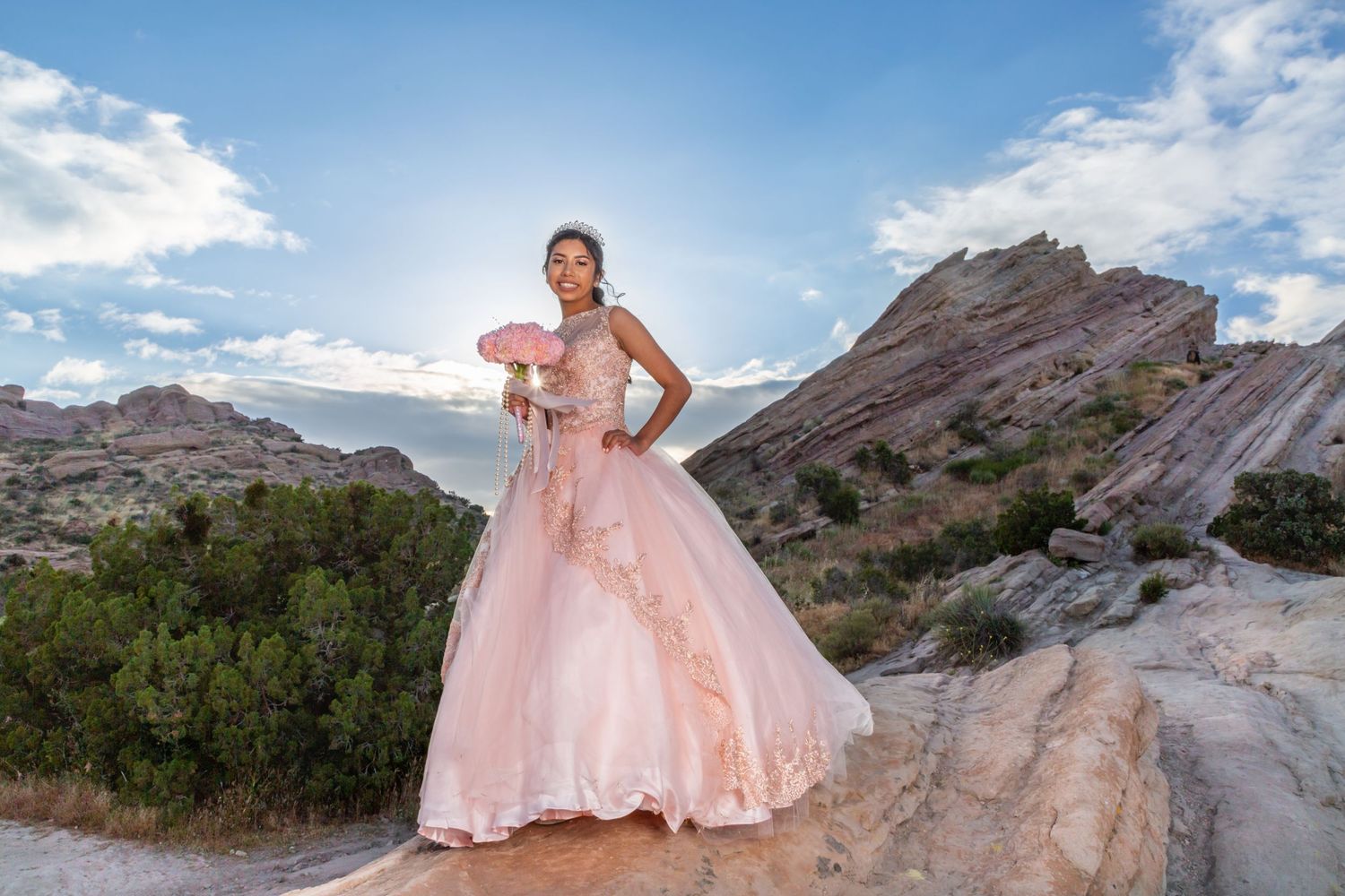A pink ball gown dress flows gracefully against the dramatic backdrop of rocky desert mountains under a blue sky.