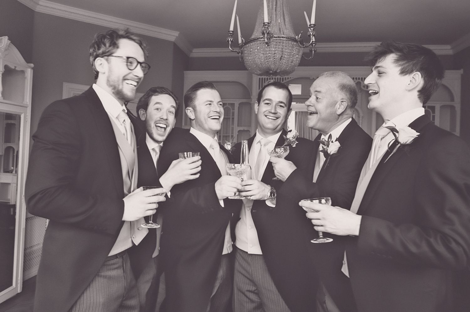 Group of groomsmen laughing and sharing drinks at indoor wedding celebration.