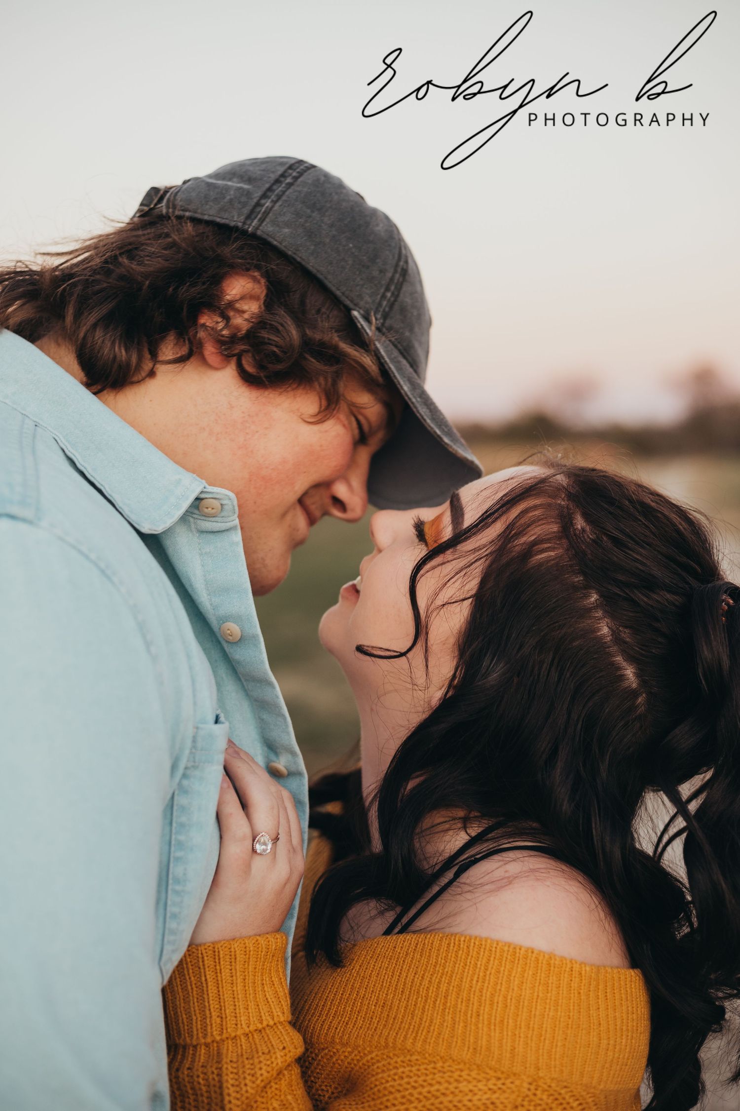 A romantic couple in casual attire sharing an intimate moment at sunset, with a photographer's watermark visible.