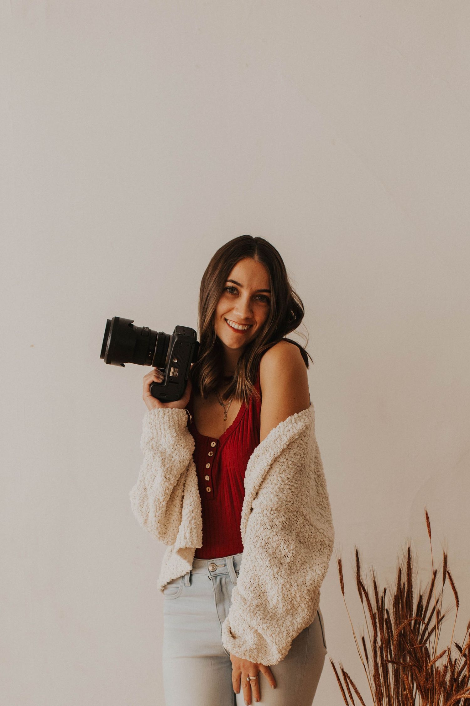 Photographer in red top and fuzzy cardigan poses casually with camera against neutral background.