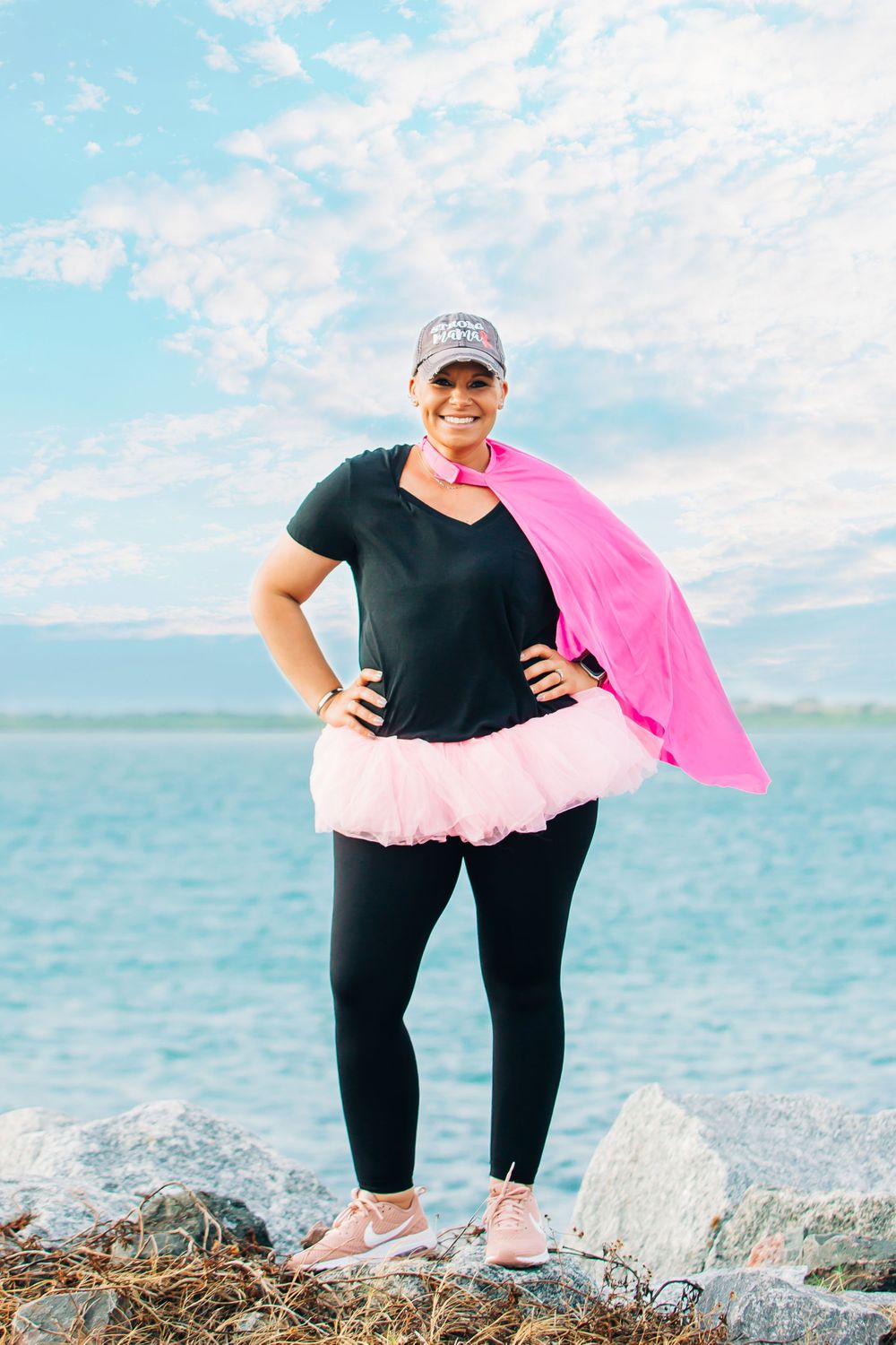 Person wearing black athletic wear and pink tutu poses confidently by the ocean.