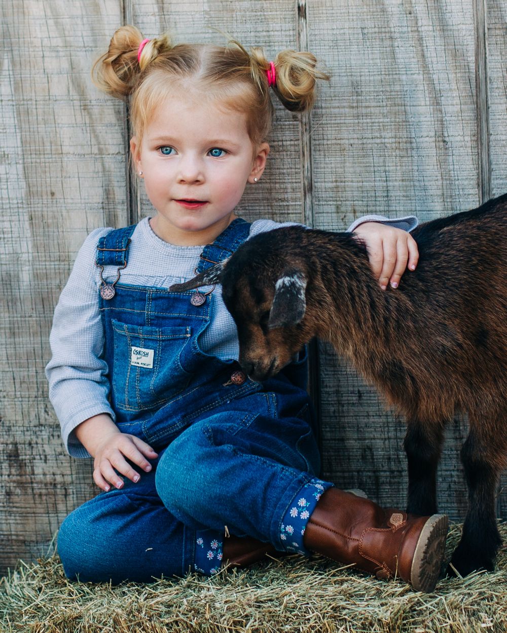 Young child in denim overalls sits with a small brown goat against a rustic wooden wall.