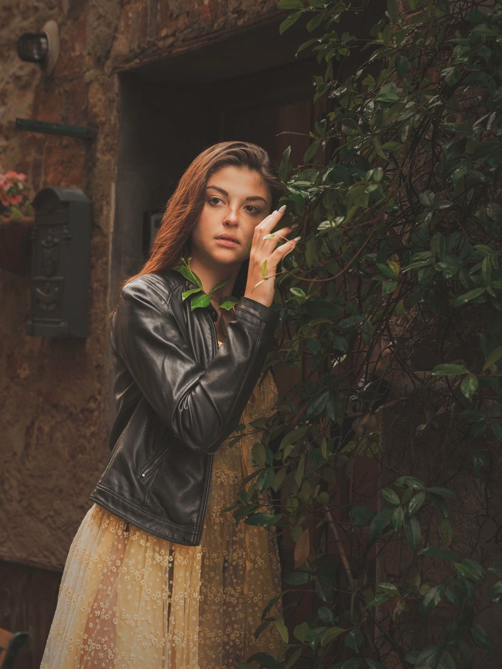 A model in a leather jacket and floral dress poses against a rustic stone wall with ivy.