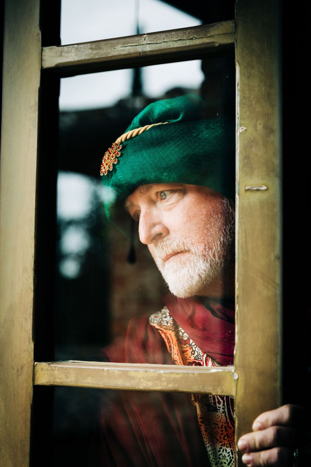 A person in medieval costume and green hat peering through a window frame.