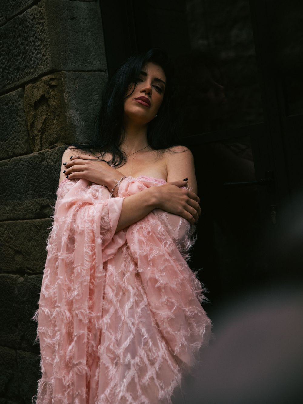 Moody portrait with soft pink lace dress against stone wall.