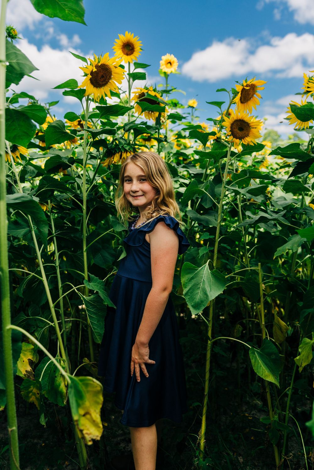 A portrait of someone in a navy dress standing in a field of towering sunflowers.